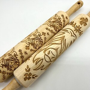 May include: Two wooden rolling pins with engraved designs of Highland Cow faces and floral patterns. The rolling pins are made of natural wood and have a light brown color.