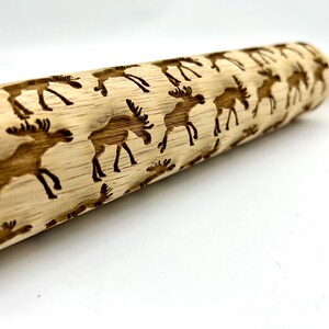May include: A wooden rolling pin with a repeating pattern of moose silhouettes engraved into the surface. The rolling pin is light brown in color and has a natural wood finish.