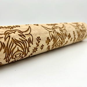 May include: A wooden rolling pin with a carved design of Highland cows and floral accents. The rolling pin is made of natural wood and has a light brown color.