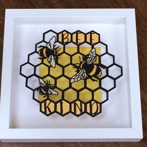 May include: A framed paper art piece with a honeycomb design and three bees. The bees are black and yellow and the honeycomb is black. The background is yellow and the text "Bee Kind" is written in black letters.