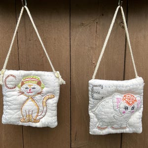 May include: Two small, white quilted bags with embroidered animal designs. One features a cat with the letter "C", the other an elephant with the letter "E". Both have cream-colored rope straps and are hanging against a wooden background.