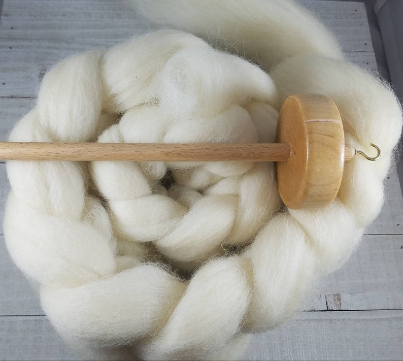 Puede incluir: Un torno de hilar de madera con una lana blanca. El torno tiene un huso de madera y un huso de madera. El huso tiene un gancho de metal.