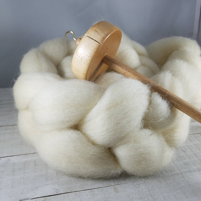 Puede incluir: Una rueca de madera con una bola de lana blanca y esponjosa. La rueca est&aacute; hecha de madera marr&oacute;n claro y tiene un peque&ntilde;o gancho de metal en la parte superior.