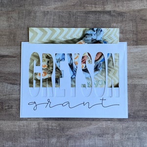 May include: White card with the name Greyson Grant cut out of the card. A baby is visible through the cut out letters.