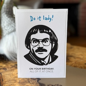 May include: A white greeting card with a black and white illustration of a man wearing glasses and a mustache. The card has the text "Do it lady!" in blue and "ON YOUR BIRTHDAY. ALL OF IT. AT ONCE." in black.