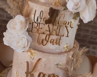 May include: A three-tiered cake decorated with white roses, dried pampas grass, and gold leaf. The cake is frosted in a light brown color and has a sign that reads "We can barely wait" and "Kam".