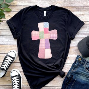 以下が含まれることがあります： ピンク、紫、緑の色合いのパッチワーククロスデザインが特徴の黒いTシャツ。十字架は様々な生地パターンで作られています。シャツは黒いスニーカーとブルージーンズと組み合わせています。