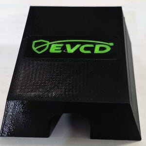 May include: A black, wedge-shaped object with a textured surface and a central cutout at the base. The top features a recessed rectangular area containing the bright green text EVCD, accompanied by a shield icon to the left of the letters.