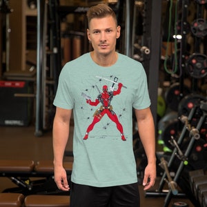 K&ouml;nnte beinhalten: Ein hellblaues T-Shirt mit einem Grafikdruck von Deadpool in einem rot-schwarzen Anzug, der ein Schwert und eine Pistole h&auml;lt. Der Hintergrund ist eine rissige Wand mit Spinnweben.