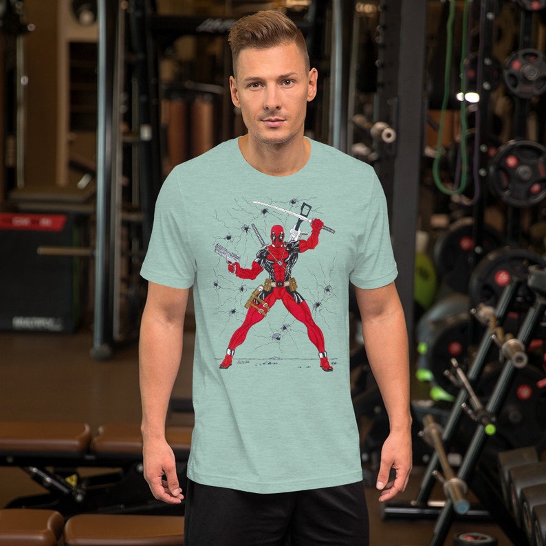 K&ouml;nnte beinhalten: Ein hellblaues T-Shirt mit einem Grafikdruck von Deadpool in einem rot-schwarzen Anzug, der ein Schwert und eine Pistole h&auml;lt. Der Hintergrund ist eine rissige Wand mit Spinnweben.