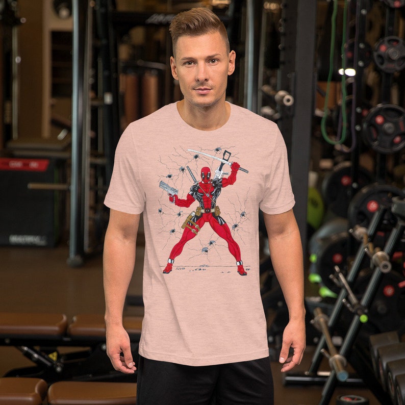 K&ouml;nnte beinhalten: Ein pinkfarbenes T-Shirt mit einem Aufdruck von Deadpool, der in einer rissigen Wand mit Einschussl&ouml;chern steht und Waffen h&auml;lt. Der Aufdruck ist in Rot, Schwarz und Wei&szlig;.