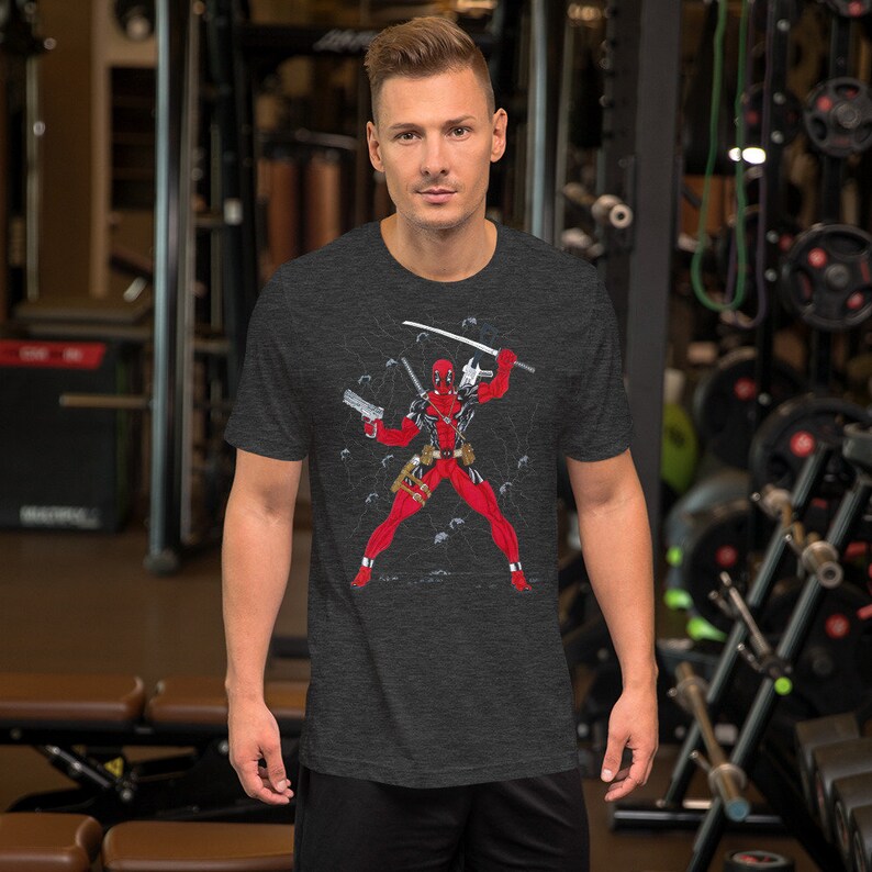 K&ouml;nnte beinhalten: Ein dunkel-helles graues T-Shirt mit einem Grafikdruck von Deadpool in Kampfpose. Er tr&auml;gt seinen charakteristischen rot-schwarzen Anzug und h&auml;lt ein Katana und eine Pistole. Die Grafik ist von einem netzartigen Muster umgeben.