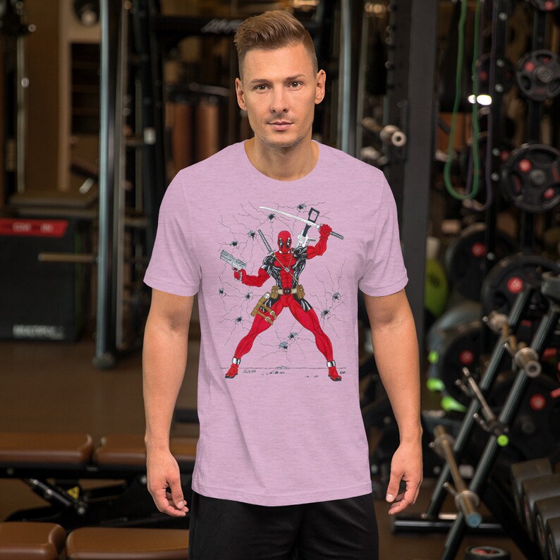 K&ouml;nnte beinhalten: Ein pinkfarbenes T-Shirt mit einem Aufdruck von Deadpool, der in einer rissigen Wand mit Waffen und einem Spinnennetz-Design steht.