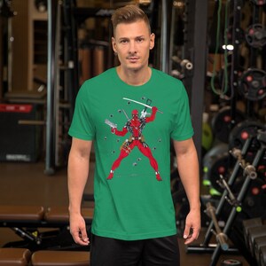 K&ouml;nnte beinhalten: Ein gr&uuml;nes T-Shirt mit einem Aufdruck von Deadpool in Kampfpose. Er tr&auml;gt seinen charakteristischen rot-schwarzen Anzug und h&auml;lt ein Katana-Schwert. Kugeln fliegen um ihn herum.