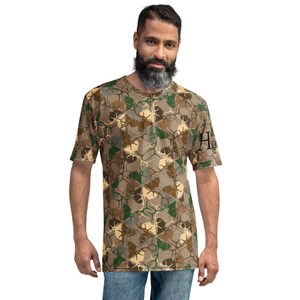 Peut inclure: Un t-shirt à imprimé camouflage avec un motif brun, vert et beige. Le t-shirt a un col rond et des manches courtes. Le texte "Home" est imprimé sur la manche droite.