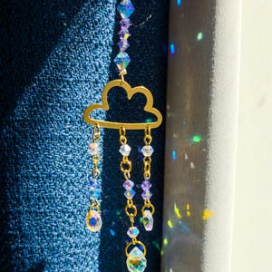 May include: A gold-toned metal cloud ornament featuring three hanging chains adorned with iridescent purple and clear faceted crystal beads. The top section includes a vertical strand of matching crystals, creating a light-catching suncatcher design.