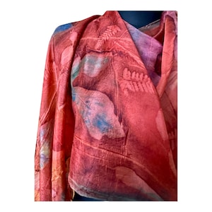 Pode incluir: Um lenço leve com uma cor base vibrante em vermelho e laranja, apresentando impressões botânicas de folhas e fetos em tons mais escuros de vermelho, roxo e azul. O tecido tem uma textura macia e drapeada com padrões de plantas.
