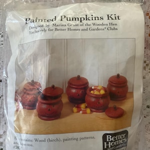 以下が含まれることがあります： Marina Grant デザインの「Painted Pumpkins Kit」。ジャック・オー・ランタンの顔が描かれた木製のカボチャの瓶が写っています。キットには木材、塗装パターン、説明書が含まれています。キャンディーコーンが入っている瓶もあります。