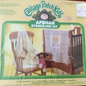 May include: Cabbage Patch Kids Afghan Stencilling Kit. The kit includes a stencil, brush, paint, and instructions. The image shows a finished afghan with a Cabbage Patch Kids design, a doll, and a wooden rocking chair. The kit is easy to crochet.