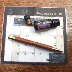 Könnte beinhalten: Ein brauner Holzstift mit goldenen Akzenten liegt auf einem Kalender mit dem hervorgehobenen Monat Februar 2019. Zwei kleine Flaschen ätherisches Öl befinden sich ebenfalls auf dem Kalender. Eine Flasche ist mit "dōTERRA Lavender" beschriftet.