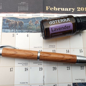Könnte beinhalten: Eine Nahaufnahme eines Februar 2019 Kalenders mit einem Holzstift und einer Flasche dōTERRA Lavendel-Ätherisches Öl. Die Flasche ist mit dem Markennamen und dem Namen des Öls "Lavender" beschriftet. Der Kalender zeigt die Daten 2, 3, 4, 5, 8, 9, 13, 14, 15, 16, 17, 18, 19, 20, 21, 22 und 23.