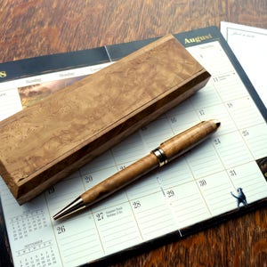 Puede incluir: Un bolígrafo de madera y una caja de madera sobre un calendario con el mes de agosto. El bolígrafo tiene una banda dorada y una punta dorada.