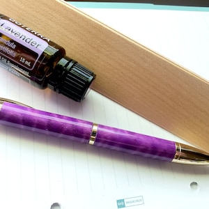 Könnte beinhalten: Ein lila-goldener Stift mit einer schwarzen und braunen Flasche mit Lavendel-Ätherischem Öl, beschriftet mit "Lavender Lavandula angustifolia 15 ml Essential Oil Supplement".