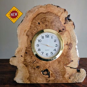 May include: A handcrafted wooden clock with a gold-tone bezel and white clock face. The clock is set in a unique, natural wood slab with a "NEW" sticker in the upper left corner. The wood has a light brown color with darker brown patterns.