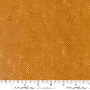 May include: A close-up of a fabric with a speckled pattern. The fabric is a warm, mustard yellow colour with tiny white dots scattered throughout. The fabric has a subtle texture, giving it a slightly rough feel.