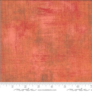 May include: A close-up of a textured fabric with a mottled orange and brown colour scheme. The fabric has a subtle brushed look and a slightly distressed appearance. The fabric is marked with a ruler and the word "moda" in a stylized font.