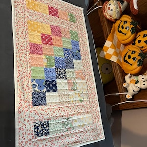 May include: A handmade patchwork quilt with a floral border. The quilt features a grid pattern of colorful squares in shades of yellow, pink, blue, and green. The border is a white fabric with a red floral pattern.