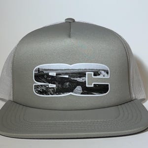 May include: A gray and white trucker hat with a black and white image of a coastal scene and the letters "SC" in the center.