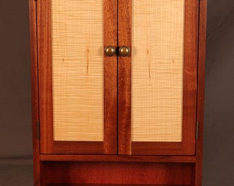 May include: A wall-mounted wooden cabinet featuring two doors with light-coloured, horizontally grained wood panels framed by dark reddish-brown wood. The cabinet includes two round brass-toned knobs and an open storage shelf at the bottom.