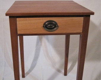 May include: A wooden side table featuring a square top and four straight legs. The table includes a single drawer with a lighter wood front panel and a centred, oval-shaped, dark metal drop handle. The frame and legs are made of a darker wood.