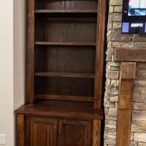 May include: A dark brown wooden bookcase with a cabinet base and open shelves. The bookcase has a traditional design with a curved top and is made of solid wood. The cabinet doors have metal handles.