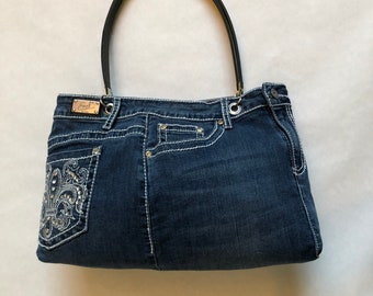 May include: A denim handbag with a black handle. The bag is made from blue jeans material and features white stitching and a decorative pocket with silver embellishments. A small metal tag is visible.