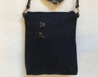 May include: A navy blue crossbody bag with a houndstooth pattern. The bag has a black leather strap, gold-coloured hardware, and two square, dark-coloured decorative elements on the front. The bag is rectangular and has a zip closure.