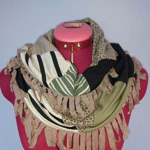 Puede incluir: Una bufanda infinita de rayas marrones, verdes, negras y blancas con flecos marrones. La bufanda está en un maniquí rosa.