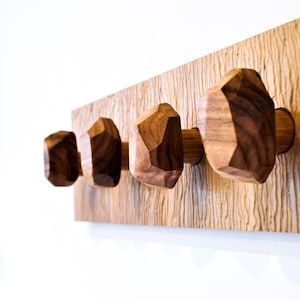 May include: A wooden wall-mounted coat rack featuring four faceted, geometric knobs. The knobs are a dark brown color and are mounted on a light-colored, textured wooden plank. The rack is designed for hanging coats, hats, or other items.