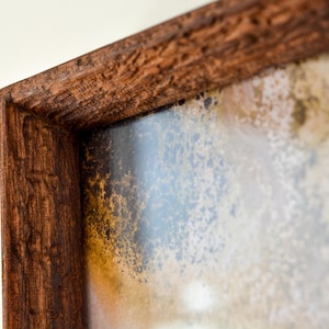 Puede incluir: Primer plano de un marco de imagen de madera con un acabado desgastado, envejecido y texturizado. El marco rodea una obra de arte con un diseño abstracto blanco, azul y marrón.
