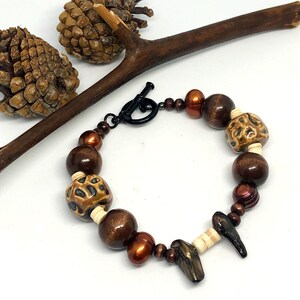 May include: A brown and white beaded bracelet with a toggle clasp. The beads are made of wood, bone, and ceramic. The bracelet has a natural, earthy feel.