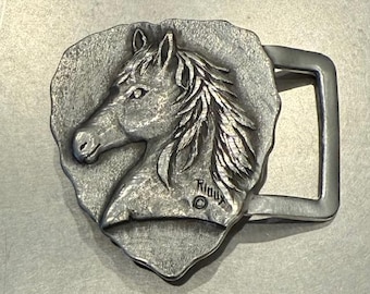 May include: A silver belt buckle with a detailed sculpted horse head design. The buckle is signed "Ridoux" on the bottom.