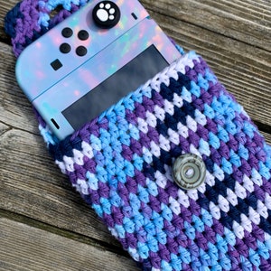 May include: A handmade crochet case in shades of blue, purple, and white, designed to hold a gaming console. The case features a button closure and a handle. The console has a holographic finish and a paw print joystick cover.