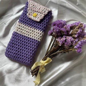 May include: A handmade crochet phone case in shades of purple, white, and light pink. The case features a button closure and a small yellow flower accent. A bouquet of dried purple flowers with a yellow ribbon sits nearby.