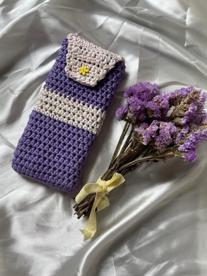 May include: A handmade crochet phone case in shades of purple, white, and light pink. The case features a button closure and a small yellow flower accent. A bouquet of dried purple flowers with a yellow ribbon sits nearby.