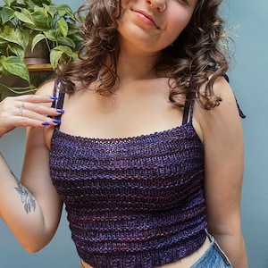 May include: A purple and pink knit crop top with thin straps. The top is made with a textured stitch and has a square neckline.