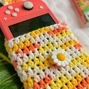 May include: A coral-colored handheld gaming device partially inserted into a handmade crochet case. The case features a daisy embellishment and a pattern of white, yellow, and orange yarn. The case is designed to protect the device.