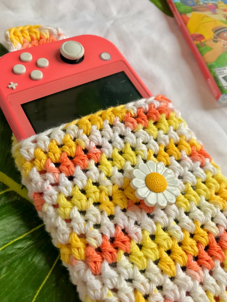 May include: A coral-colored handheld gaming device partially inserted into a handmade crochet case. The case features a daisy embellishment and a pattern of white, yellow, and orange yarn. The case is designed to protect the device.