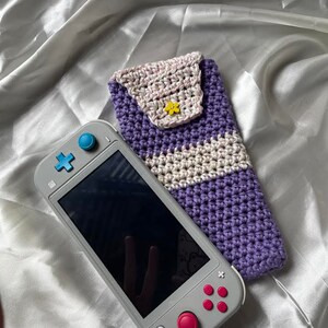 May include: A gray handheld game console next to a handmade crochet case. The case is purple and white with a small yellow flower button. The game console has a black screen and pink and blue buttons.