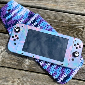 May include: A handheld gaming console with a pearlescent finish, resting on a crocheted sleeve. The console features paw print designs on the buttons and is set against a backdrop of a wooden surface. The sleeve is a mix of blue, purple, and white yarn.